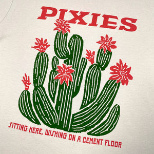 Close up of a beige T-Shirt with green and pink graphic displaying a cactus. Pink lettering reads "Pixies, sitting here, wishing on a cement floor"