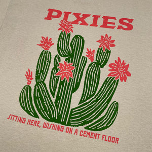 A close up of a sand t-shirt featuring a graphic of green cacti with light red flowers and the word 'PIXIES' printed in light red above them and light red text reading "Sitting here, wishing on a cement floor" below.
