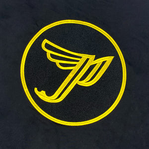 A close up of the back of a black bomber jacket with a yellow "Circle P" Pixies logo centered on the back. There is a yellow tag on the sleeve with black text reading "Remove Before Ignition."