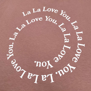 Close up of the back of a hazy Pink Crop Top against a white background with white text circling like a whirlpool reading "La La Love You." repeatedly.