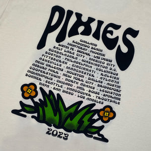 A close up of the back of a natural t shirt with black text reading "Pixies." Listed below are all 2023 Tour dates in the shape of a mushroom head. Below the list of cities is a grass and flower design with black text underneath reading "2023"